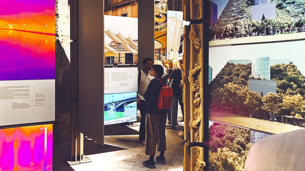 Part of "In the Fold of Shadows: Prosthetic Dialogues" -- there are lighted panels with color photos of buildings, and people are looking at them.