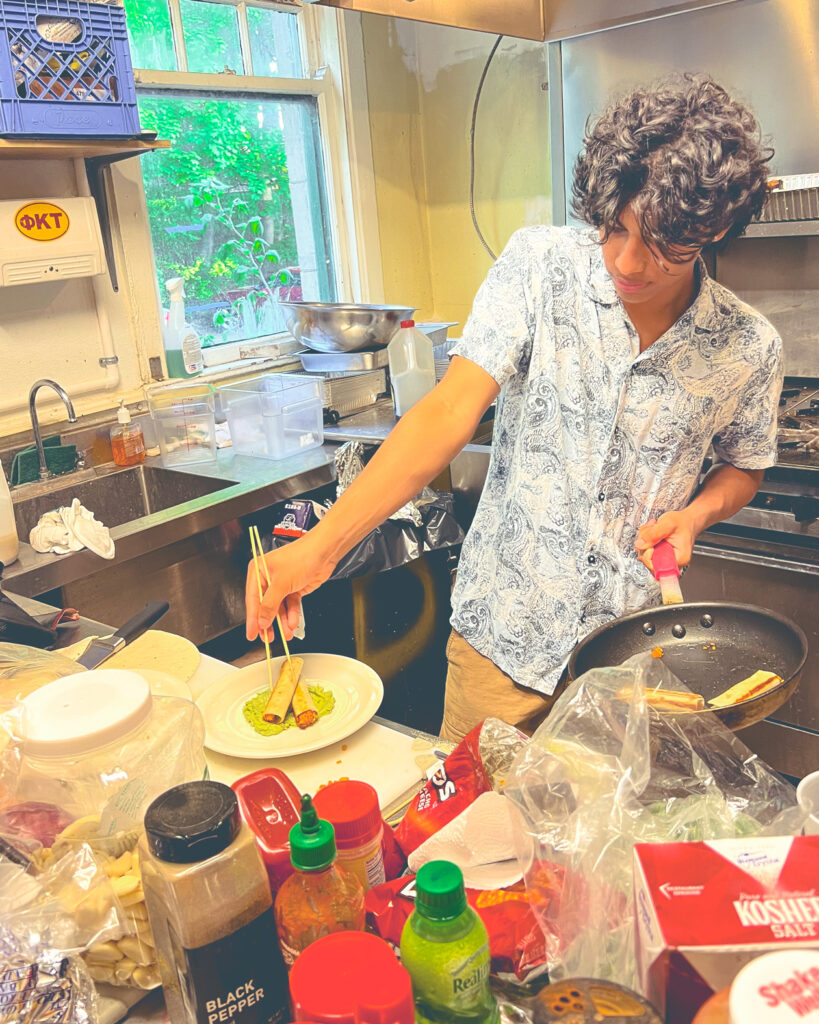 Arnav Kamulkar in his fraternity house kitchen preparing a food dish with one hand and holding a frying pan in the other.