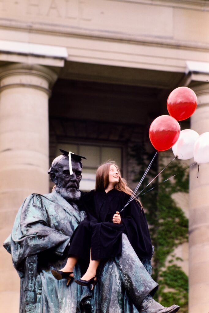 a graduating senior holding balloons sits on the lap of the A.D. White statue in 1984