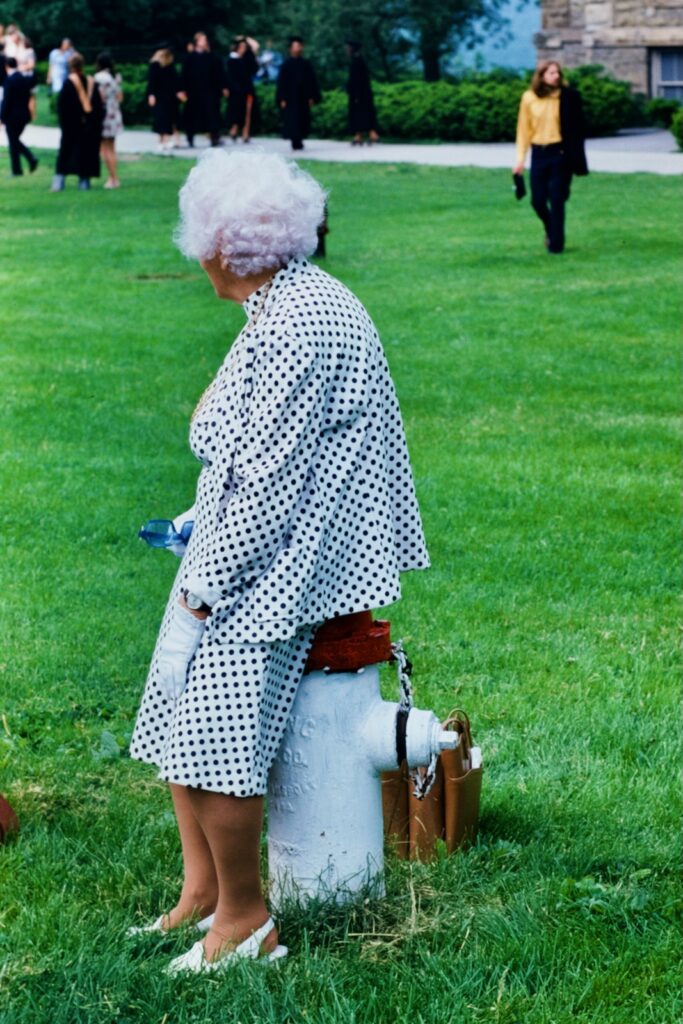 an older woman leans against a fire hydrant as graduates assemble on the Arts Quad in 1974