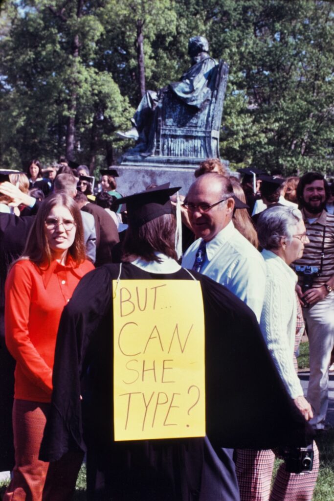 a student wears a "but .. can she type?" sign on the back of a graduation gown in 1976