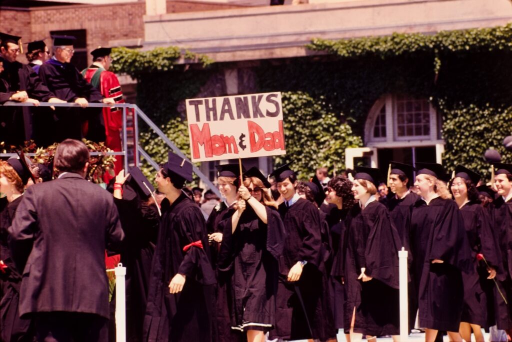 graduates processing past the review stand on the Arts Quad hold a sign stating "Thanks Mom & Dad"