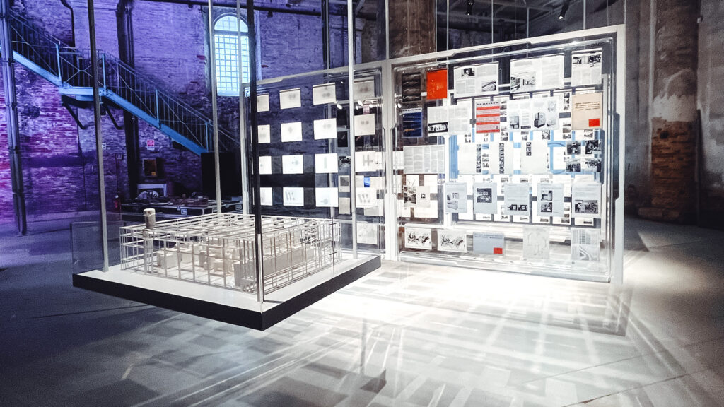 "Doxiadis’s Informational Modernism" depicts a hanging square platform with an architectural model. There are freestanding glass display cases with documents on them.