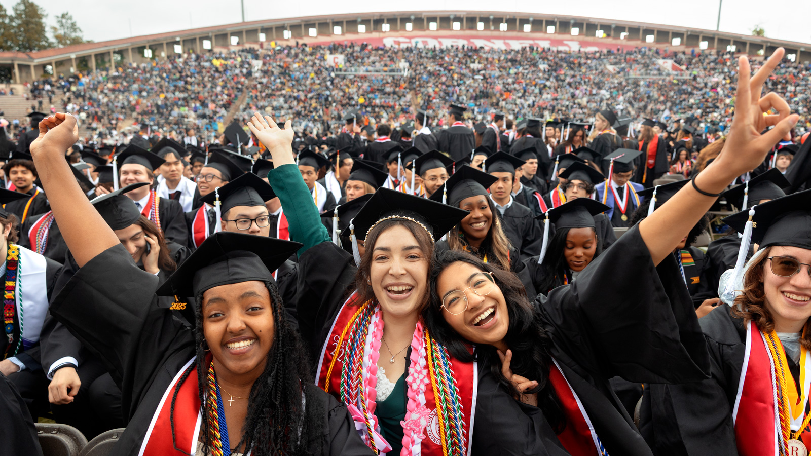Three women graduates from the class of 2025 in their academic regalia smile and wave their hands in their air during commencement at Schoellkopf Field.