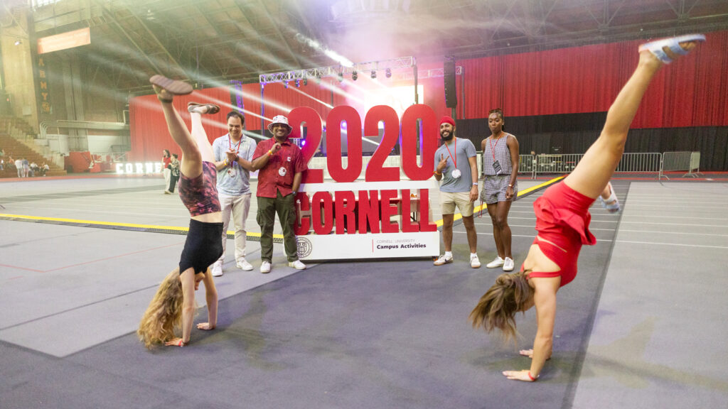 Two women do handstands in Barton Hall in front of a 2020 CORNELL sign