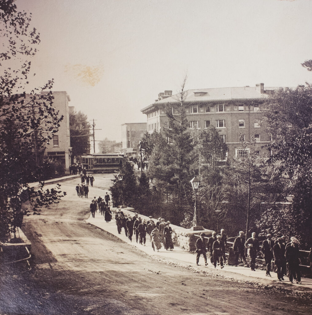 pedestrians cross the bridge in Collegetown to campus, with a trolley car and Sheldon Court in the background, early 1900s