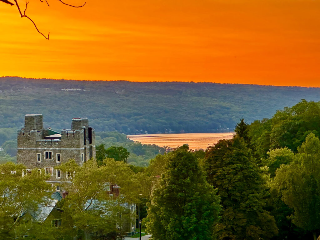 A sunset over Cayuga Lake with one of the Baker residence halls visible at left among the treetops