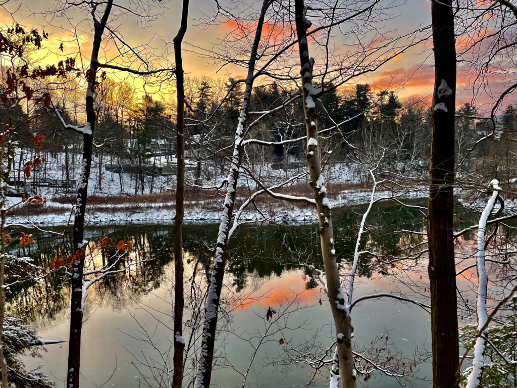 The yellow, pink, and blue sky reflect off of the water at Beebe lake during a sunset with snow-dusted trees in the foreground