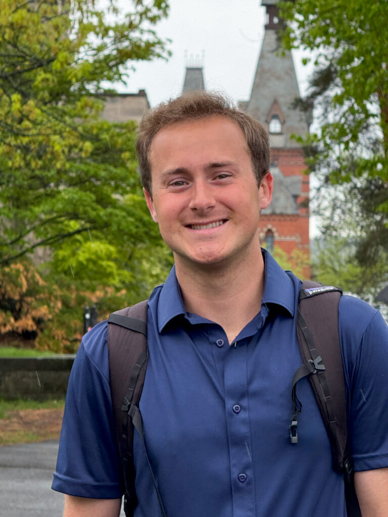 Garrett Spillerman smiles while wearing a blue collared button-down and backpack near Sage Hall