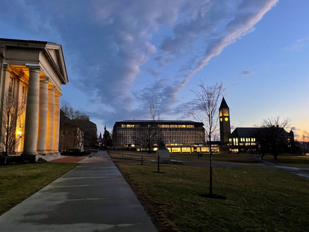 The quad, with Goldwin Smith Hall, Olin Library, McGraw Tower, and Uris Library illuminated against a blue sky and streaked clouds as the sun sets