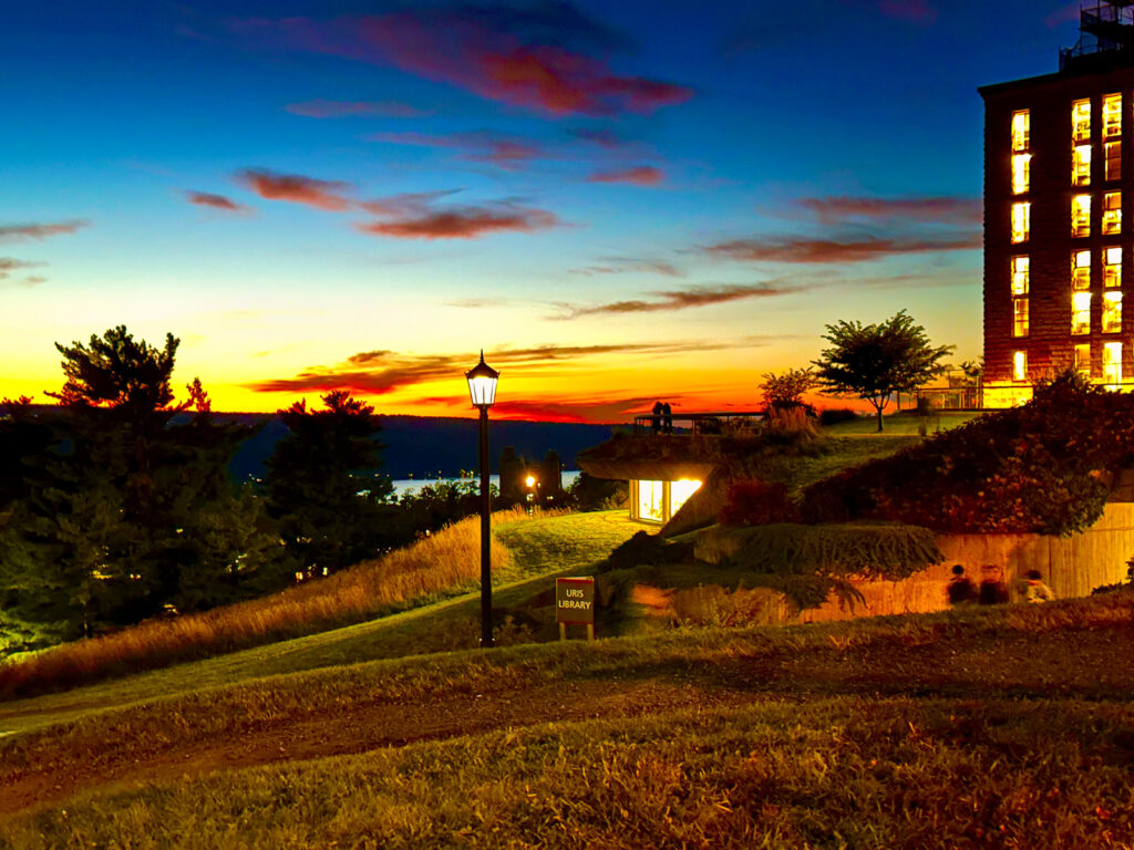 Uris Library, illuminated by its lights during sunset, with a sliver of Cayuga Lake visible in the background