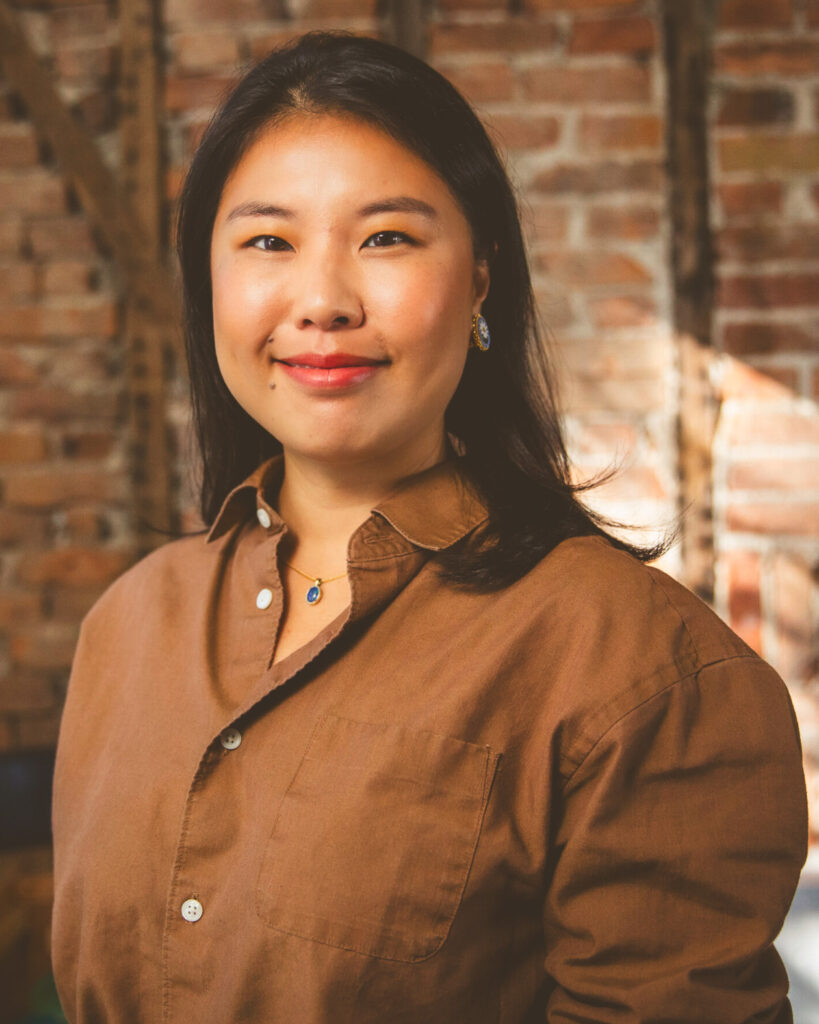 Heather Jiang smiles wearing a light brown button down standing in front of a brick wall.