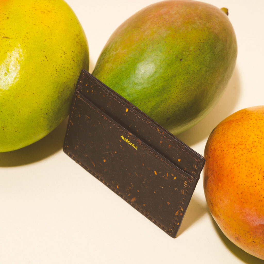 A card holder from Allegorie made from mango, surrounded by three mangos, that shows flecks of orange mango in the dark brown leather.