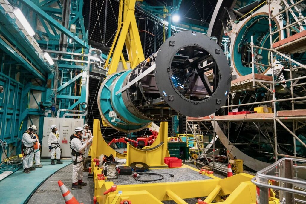 The car-sized cylindrical commissioning camera in the process of being lifted by a yellow crane. The telescope structure is visible to the left, ready for the camera's installation