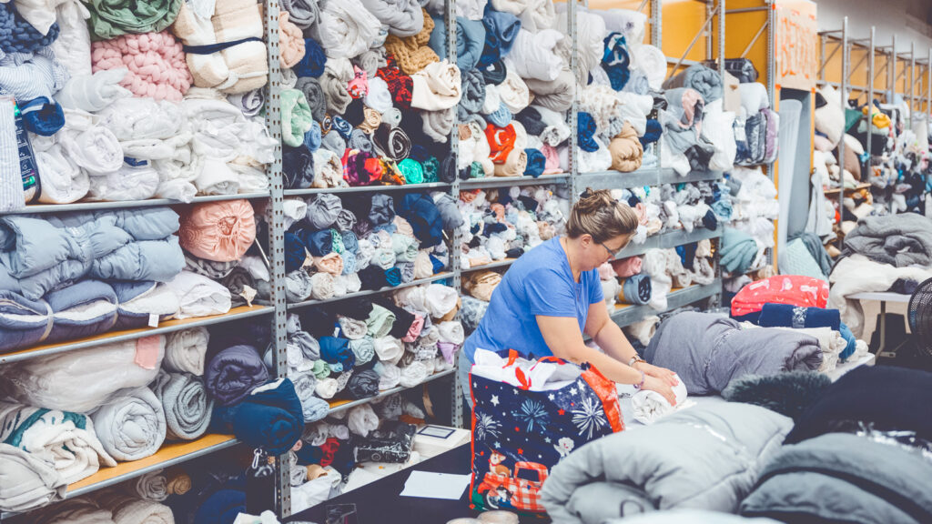A volunteer wearing a blue shirt folds blankets to add them to the packed, colorful wall of blankets behind her while preparing for the Dump and Run sale in the Ithaca Mall.