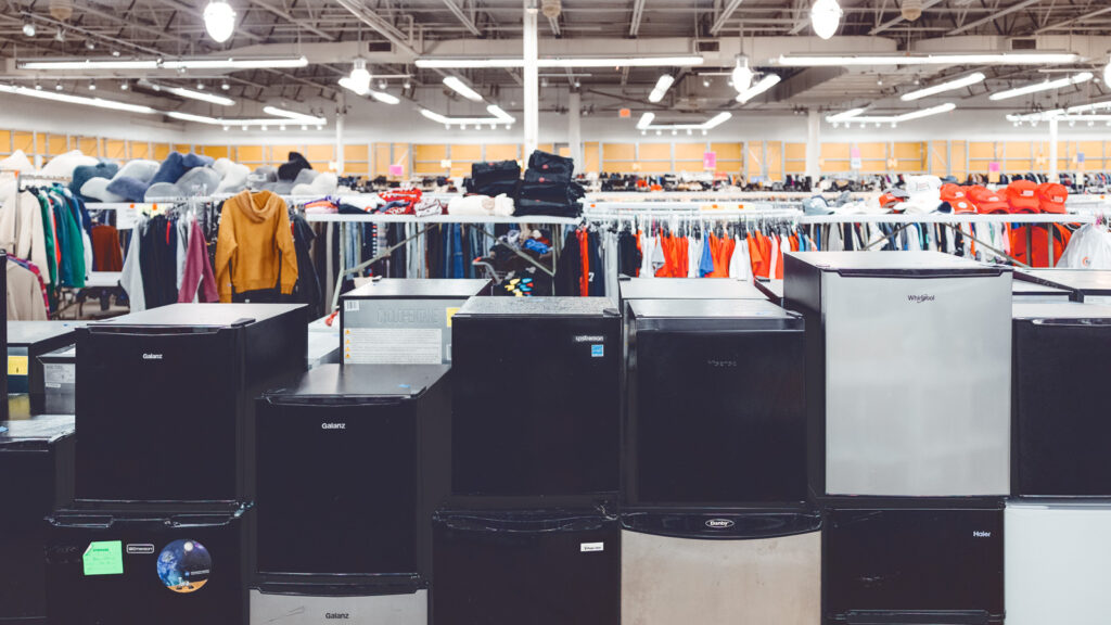 Black and gray mini fridges stacked on top of each other with racks of clothing behind the fridges at the site of where the Dump and Run sale will be in the Ithaca Mall.