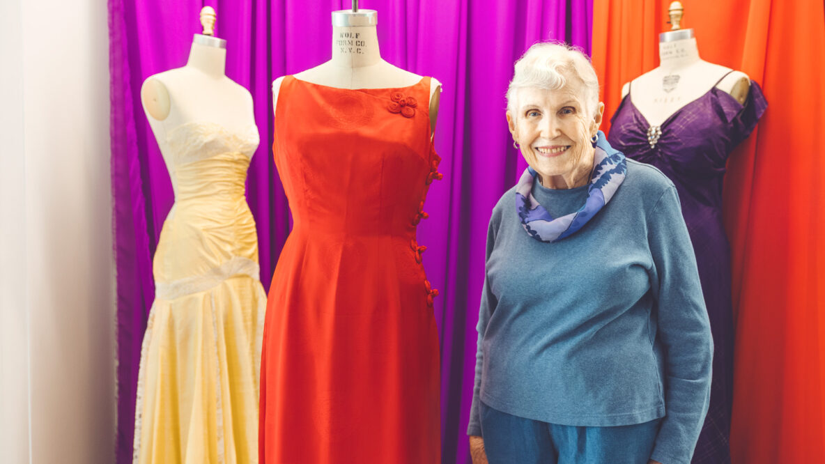 Peggy Dunlop stands next to her red dress on a mannequin and smiles.
