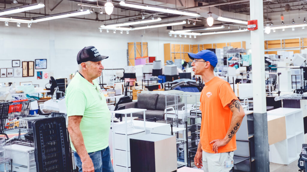 Bill Apgar, wearing a green shirt and black hat, talks with a volunteer wearing an orange shirt and blue hat while preparing for the Dump and Run sale at Ithaca mall.