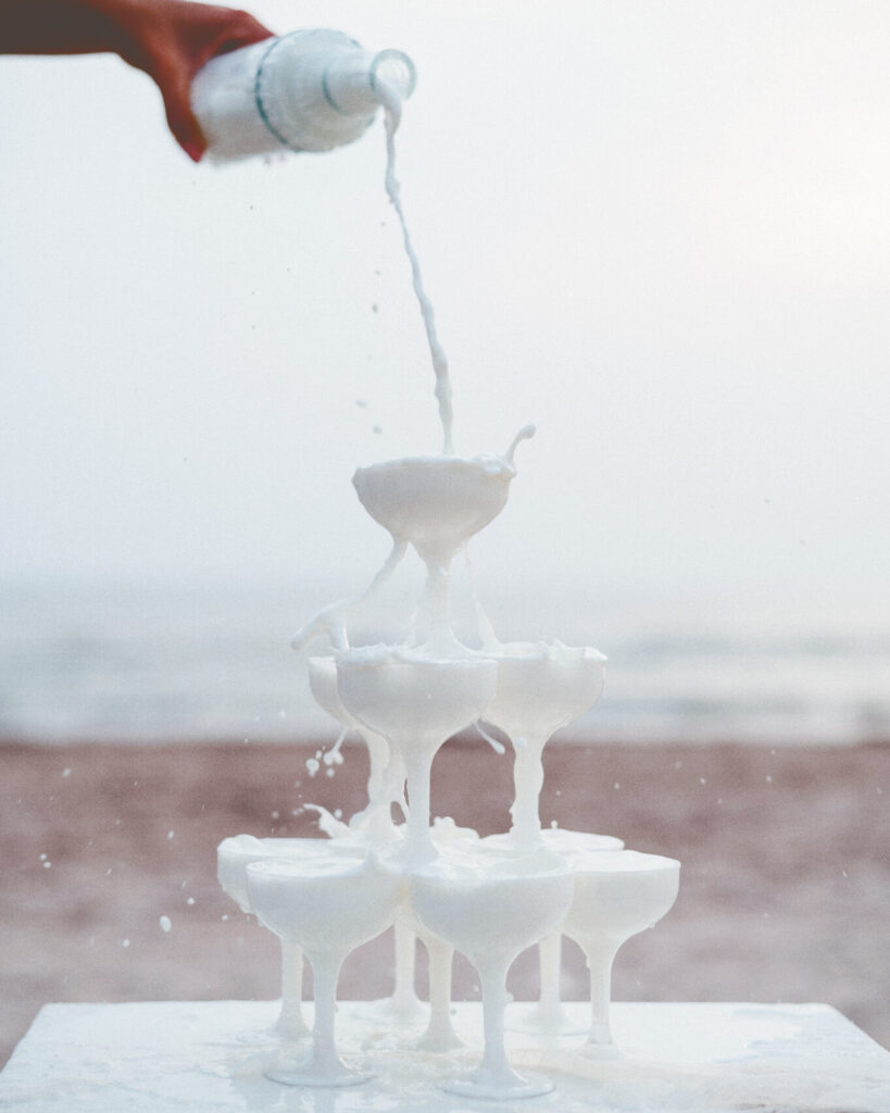 Milk is poured from a glass bottled onto an overflowing champagne tower of glasses filled with milk.