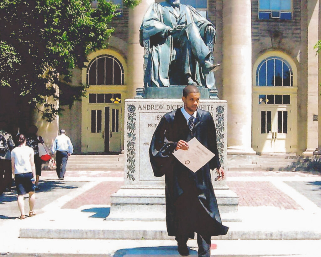 Kevin Gibson holding his diploma, wearing a graduation gown in front of the A.D. White statue