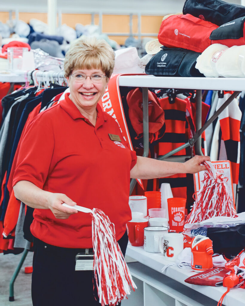 Cindy Lockwood, smiles while wearing a red Cornell collared shirt and holds two pom poms she picked up from a table of Cornell stuff for sale at Dump and Run.
