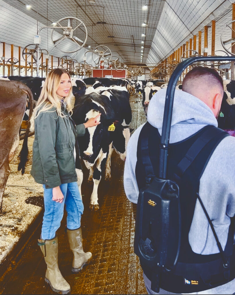 Hailey Pipher stands in a cow barn and talks while she is filmed by a camera crew.