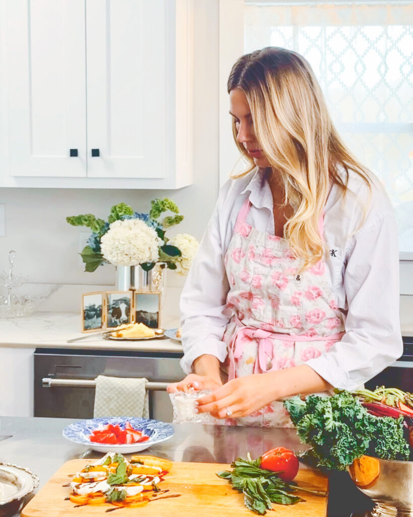 Hailey Pipher wearing an apron in the kitchen with a bowl of vegetables on the table preparing caprese.