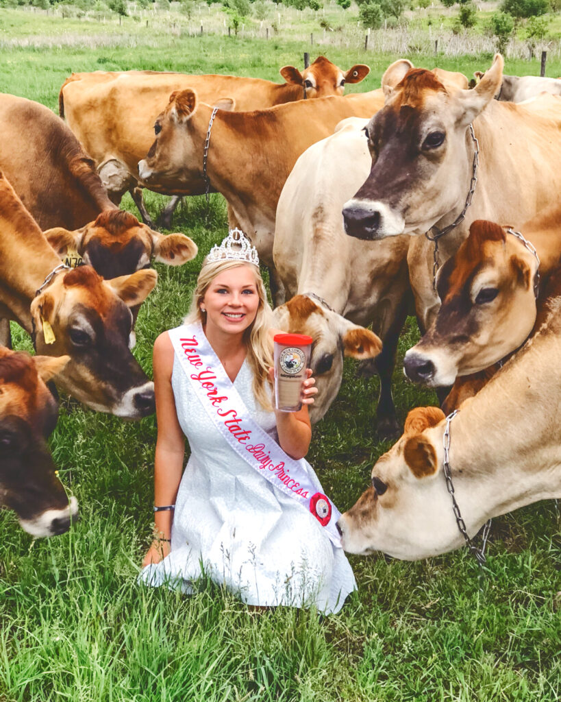 Hailey Pipher surrounded by cows in a pasture holding up a glass of milk as the New York State Dairy Princess, wearing her sash and crown.