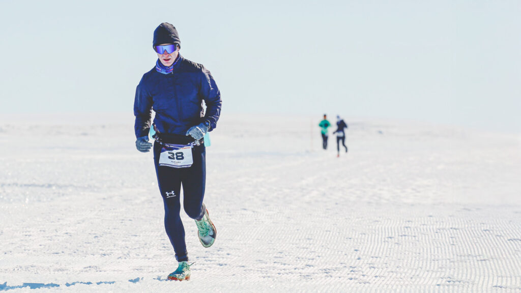 Alan Nawoj runs through the snow while wearing a hat, glasses, coat, gloves, and leggings during the Antarctica race.