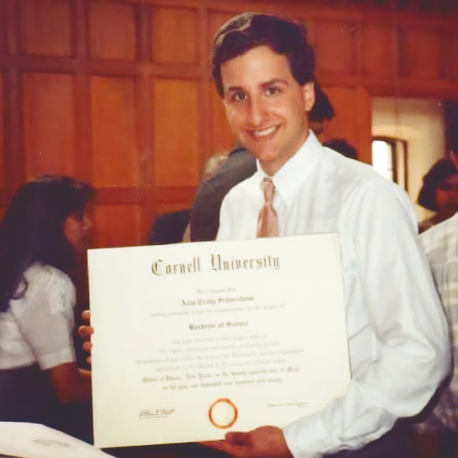 A young Alan Schussheim holding his Cornell University diploma and smiling while wearing a white dress shirt and tie