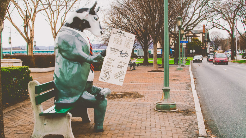A fiberglass cow sitting on a park bench, reading a newspaper