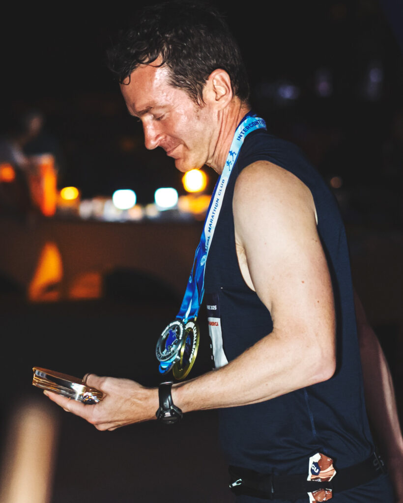Alan Nawoj looks down at a medal he has won and smiles while wearing three medals around his neck after completing the challenge in Miami.