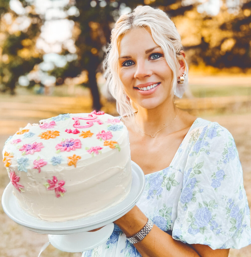 Hailey Pipher stands outside in a white and blue floral dress and smiles while holding up a white cake decorated with colorful floral icing.