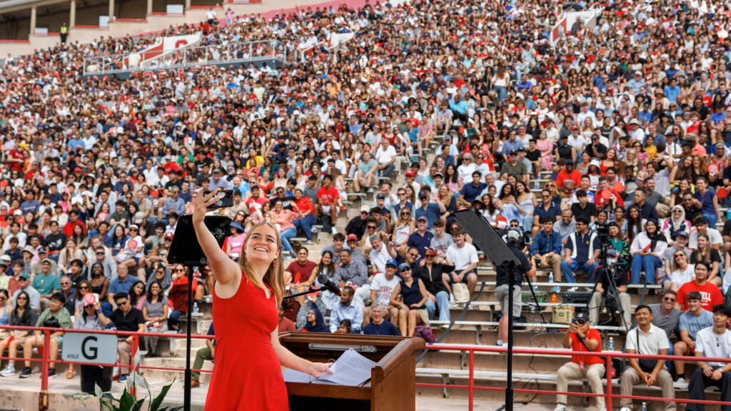 A student in a red dress smiles and holds her arm back to take a selfie on stage with the crowd behind her in the bleachers at Schoellkopf Stadium