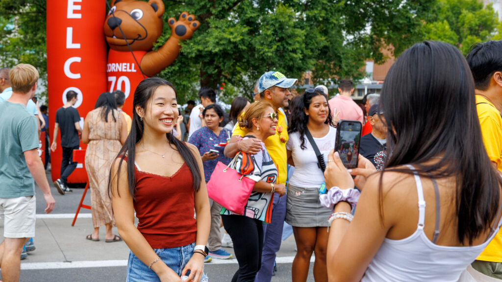 A new student smiles while wearing a red tank top while a friend takes a picture of her on her cell phone. A family poses for a photo in the background.
