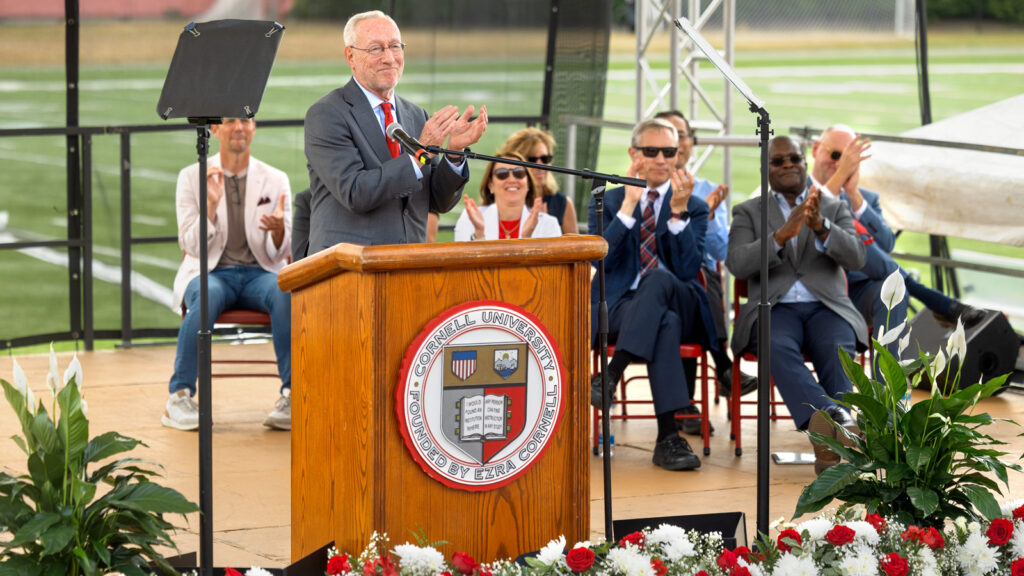 President Michael Kotlikoff wears a gray suit and red tie and claps behind the podium during new student convocation at Schoellkopf Field
