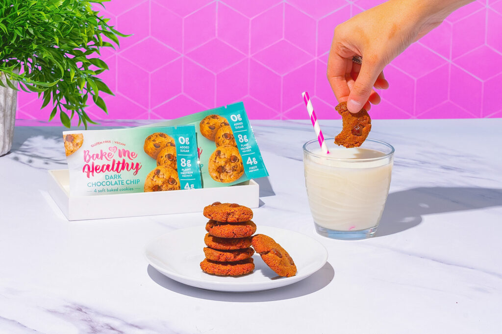 Dark chocolate chip cookies on a plate with a hand dunking a cookie into a glass of milk on the right and a Bake Me Healthy cookie package on the left.