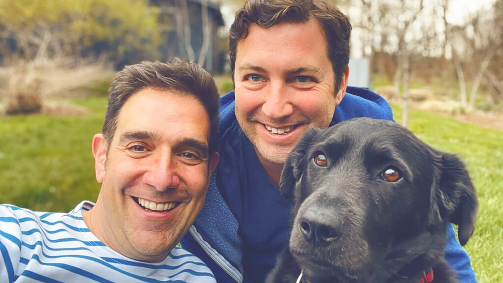 Mat Zucker and his husband pose for a selfie with their dog in their yard.