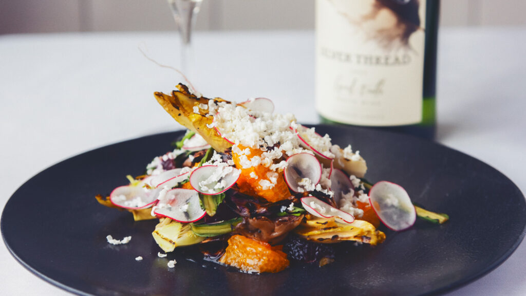 A grilled salad on a black plate paired with a bottle of wine from Silver Thread Vineyard behind the plate.
