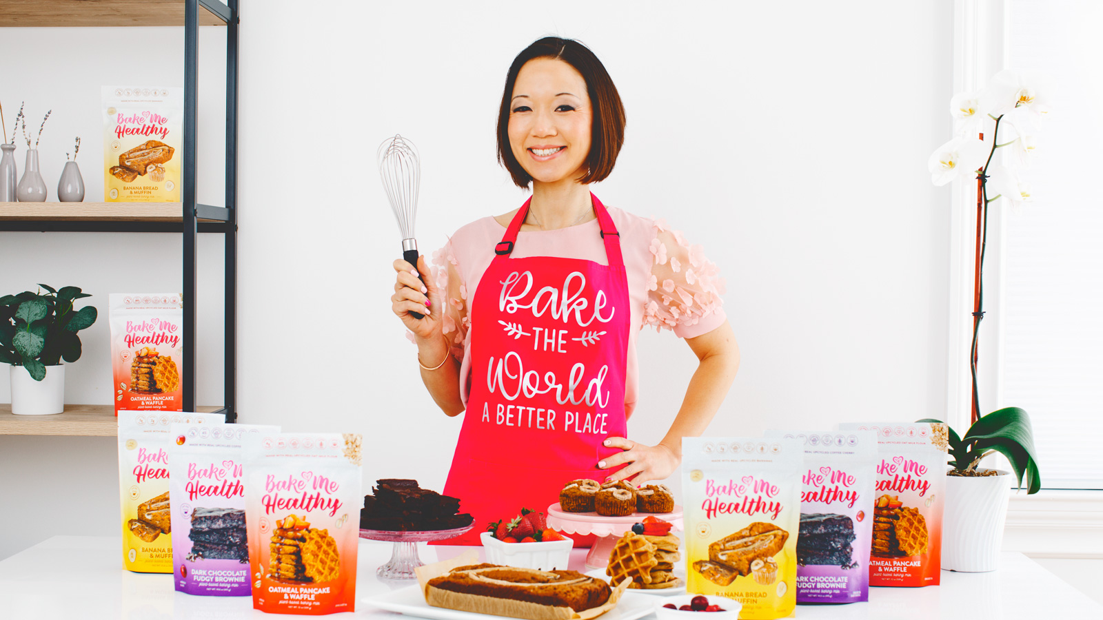 Kimberle Lau holds a whisk up and smiles in front of desserts and bags of her mixes.