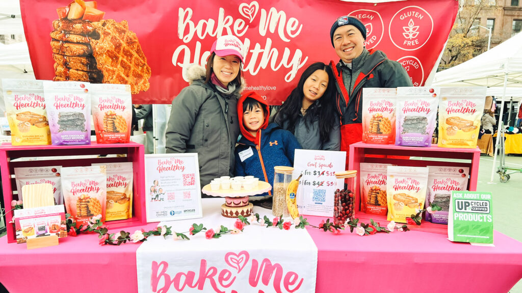 Kimberle Lau with her husband and two kids behind a pink Bake Me Healthy display table with a banner and products on display.