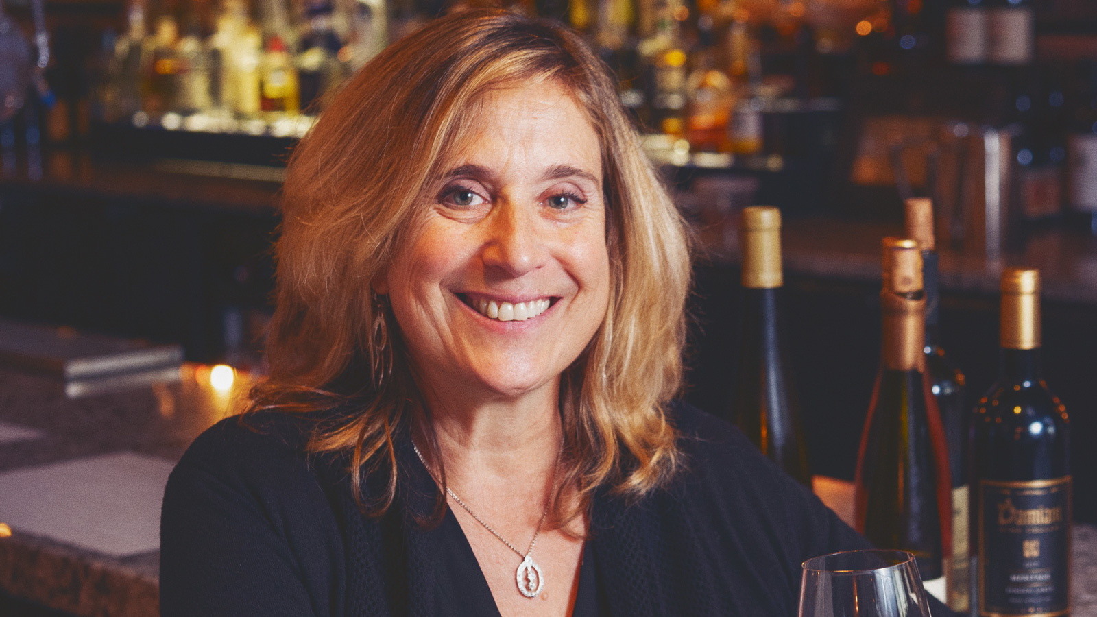Laura Winter Falk sits at a bar and smiles while holding a glass of wine.