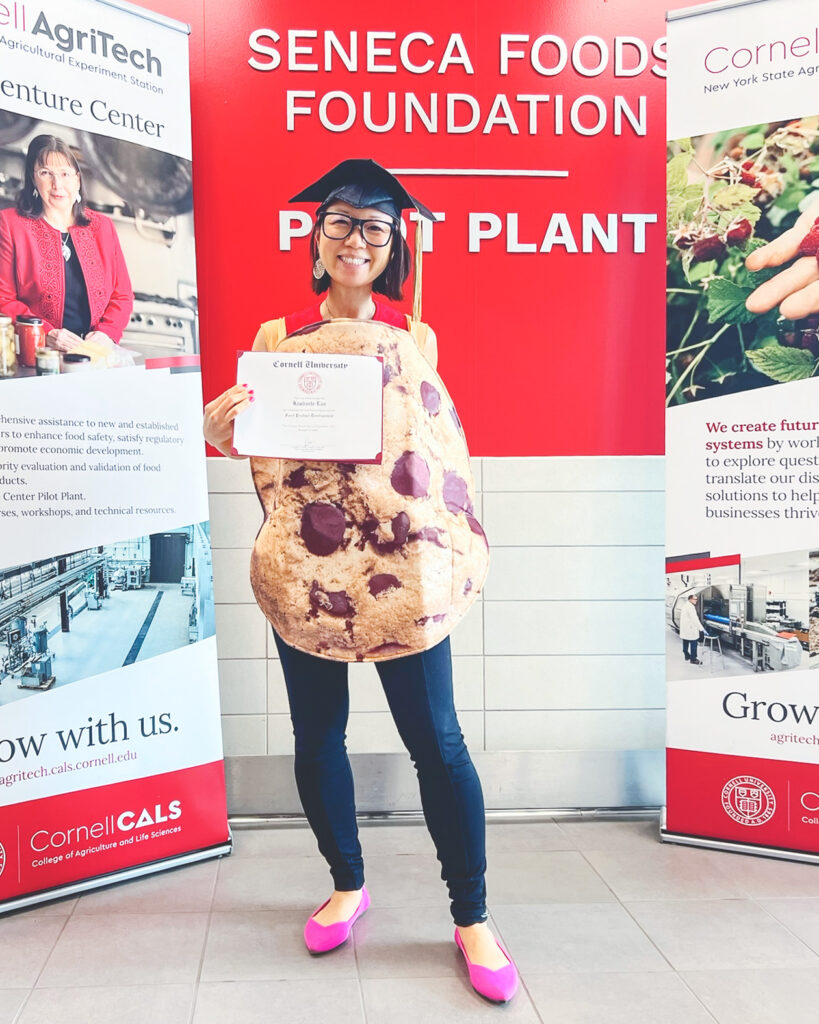 Kimberle Lau in a chocolate chip cookie costume holding her diploma and wearing her graduation cap in front of posters for CALS.