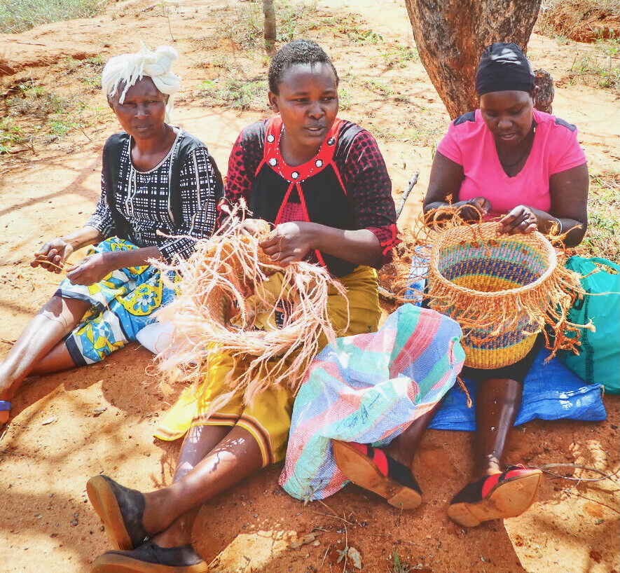 Three women in Kenya sit underneath a tree while weaving baskets.