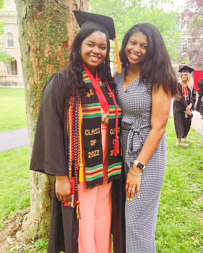 Shelby and Laura Holland stand next to each other and smile during Laura's graduation while Laura wears her cap, gown, and cords.