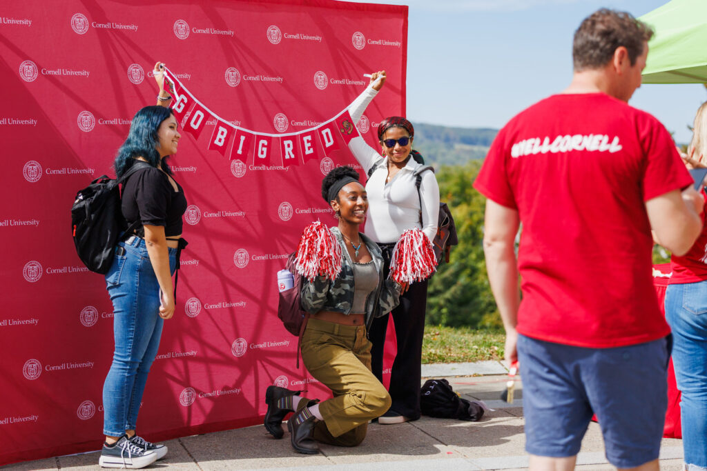Three girls pose in front of a red Cornell photo backdrop while holding a banner that says "Go Big Red" during spirit week.