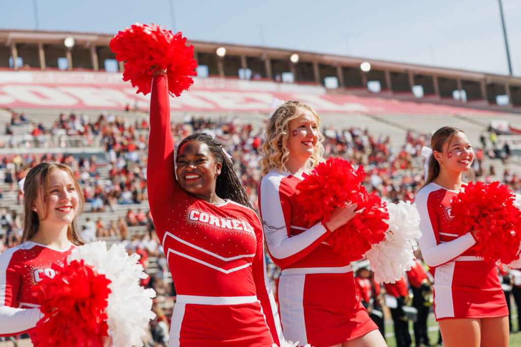 Four cheerleaders smile, cheer, and hold their pompoms while watching the homecoming football game.
