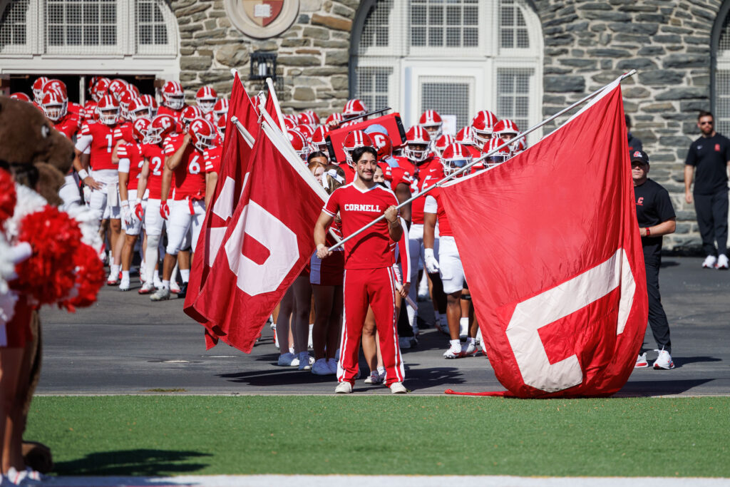 Students stand with the large red Cornell flags and wait to run onto the field while the football stands waiting behind them.