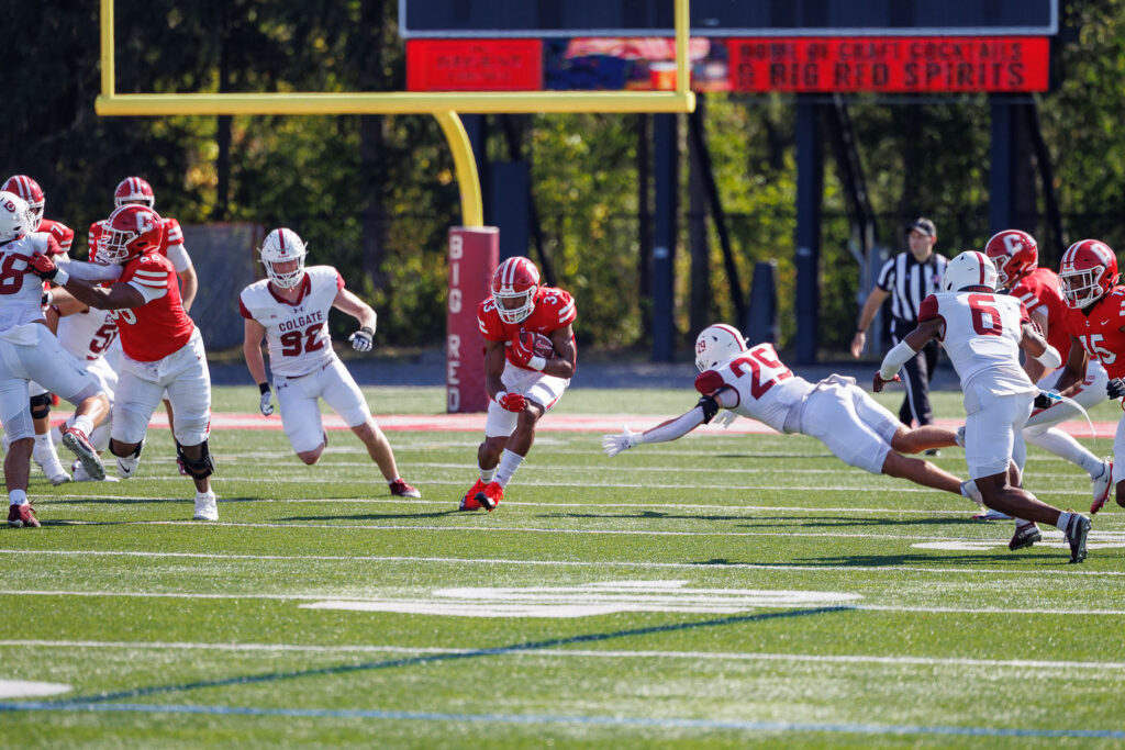 A Cornell football player runs through the middle of the field as a Colgate player dives to tackle him.