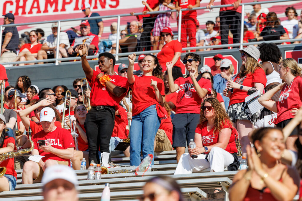 Three students wearing red stand and dance with their hands in the air on the bleachers at Schoellkopf Field during the homecoming game.